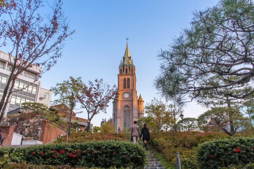 The brick Gothic bell tower and clock face of Myeongdong Cathedral, seen from the garden steps in autumn