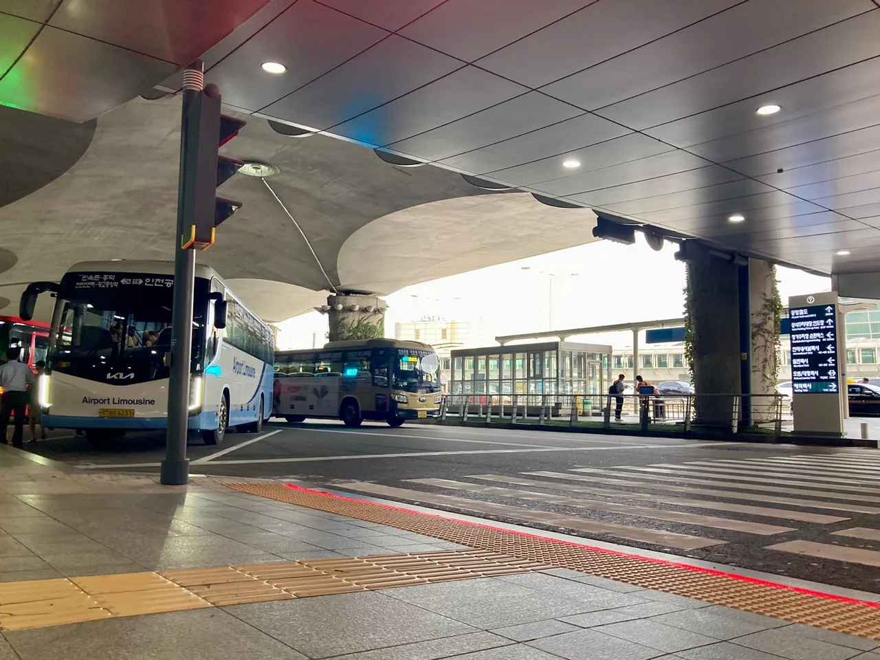 The kerbside bus boarding area on the first floor of Incheon Airport Terminal 1, with an Airport Limousine coach at the stop beside a waiting shelter and a direction sign