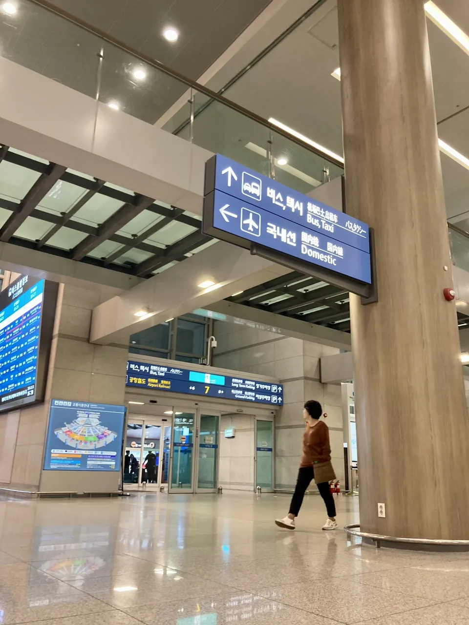 A blue direction sign in the Terminal 1 arrivals hall: an up arrow for Bus, Taxi and a left arrow for Domestic, with a board beneath it pointing to the Airport Railroad and gate 7