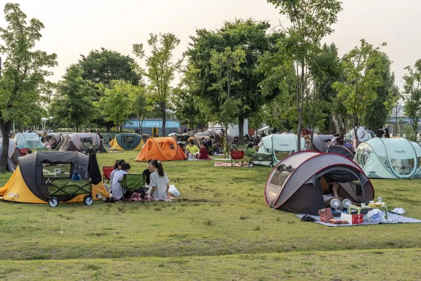 여의도한강공원 잔디밭에 텐트와 그늘막이 빼곡히 서 있고, 돗자리를 편 사람들이 삼삼오오 앉아 음식을 펼쳐 놓고 있다. 접이식 왜건과 캠핑 의자가 텐트 사이사이에 놓여 있다